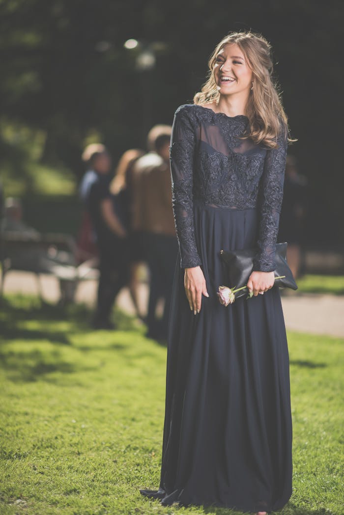 Smiling young woman in a dark evening gown holding a flower while standing outdoors.
